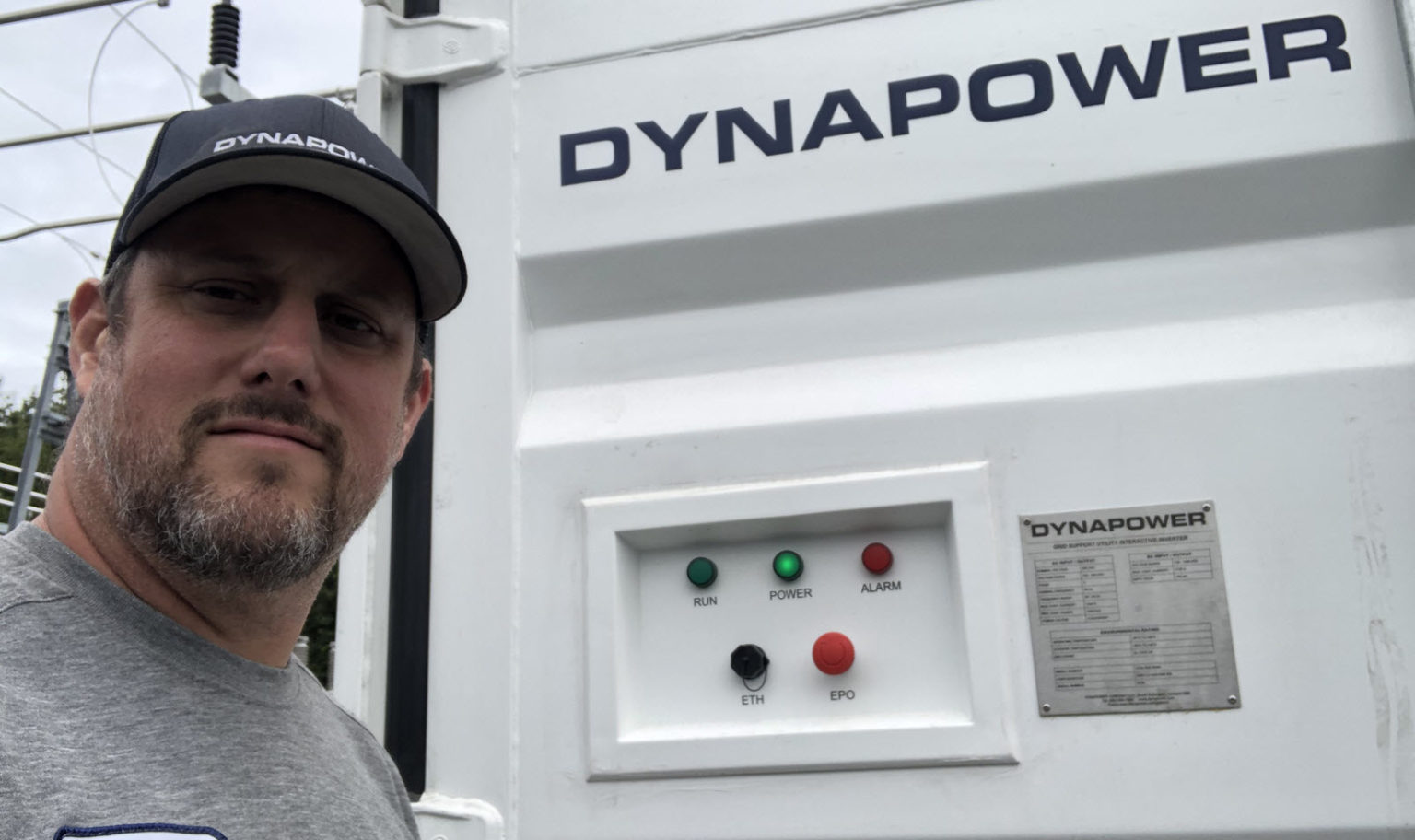 Patrick Carroll in Dynapower branded hat in front of Dynapower energy storage system