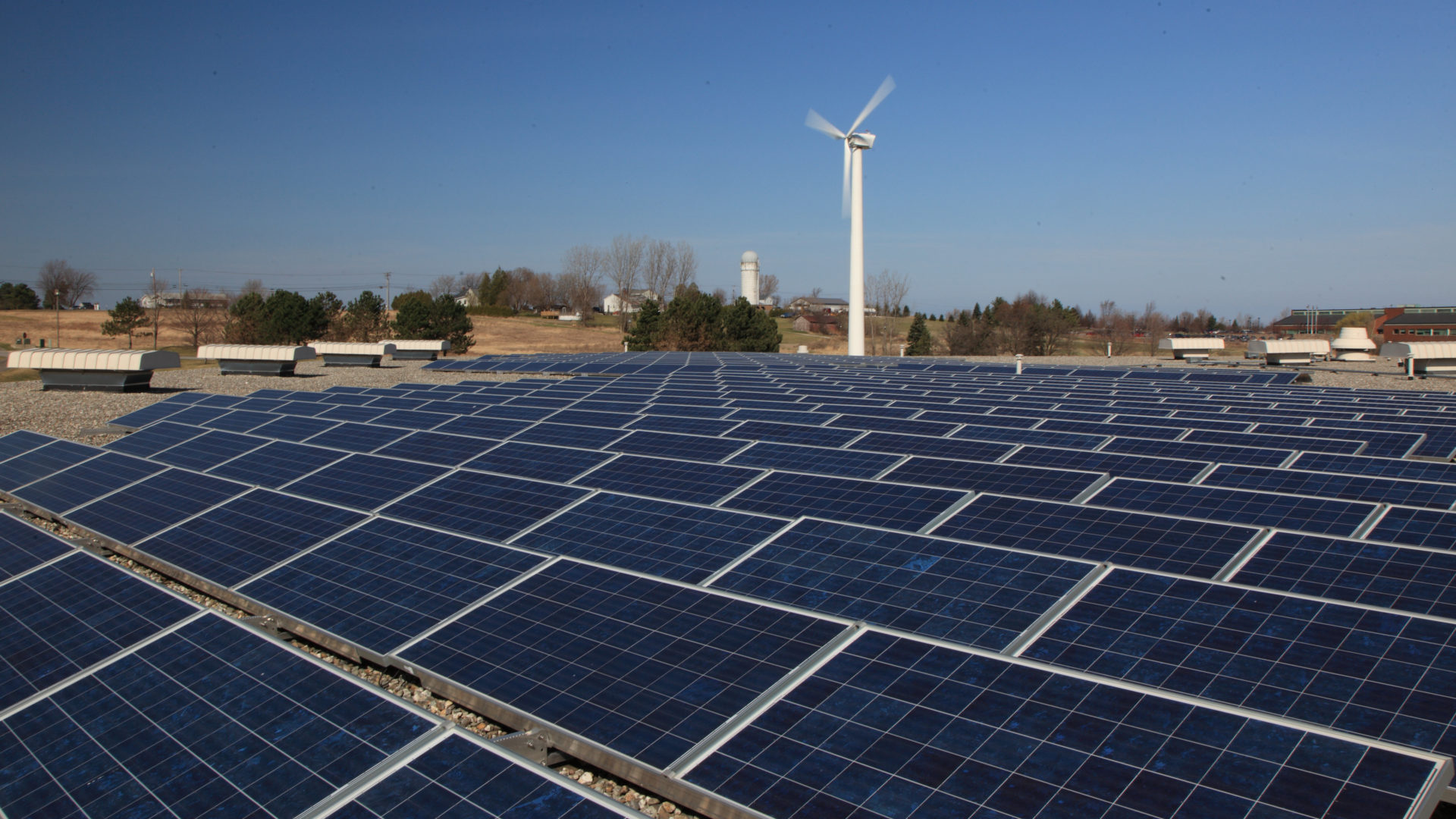 rows of solar panels with wind turbine and farm in background