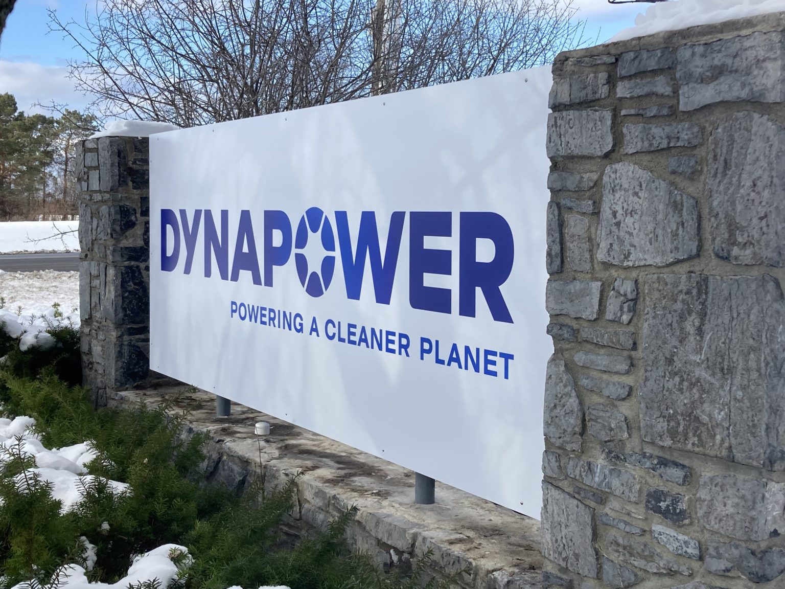 stone wall with white sign that reads "Dynapower, Powering A Cleaner Planet" in blue